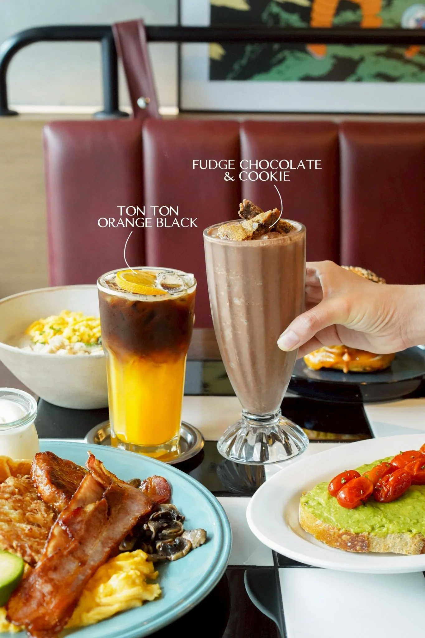 A table with breakfast food and drinks, including a bowl of scrambled eggs, bacon, mushrooms, avocado toast topped with cherry tomatoes, two beverages with labels: iced coffee labeled 'Ton Ton Orange Black' and a chocolate milkshake labeled 'Fudge Ch
