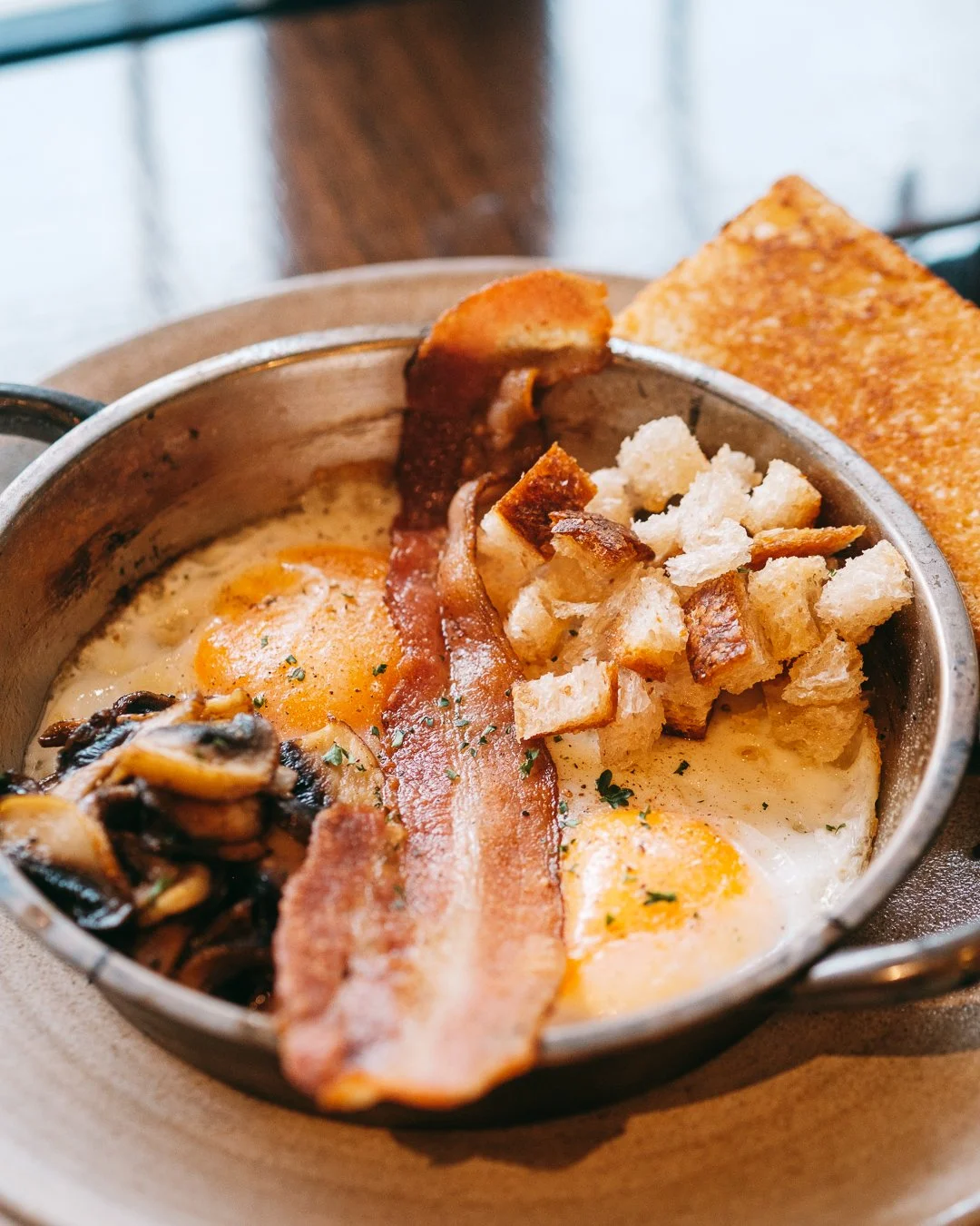 Fried eggs with bacon, sautéed mushrooms, croutons, and a slice of toasted bread served in a small skillet.