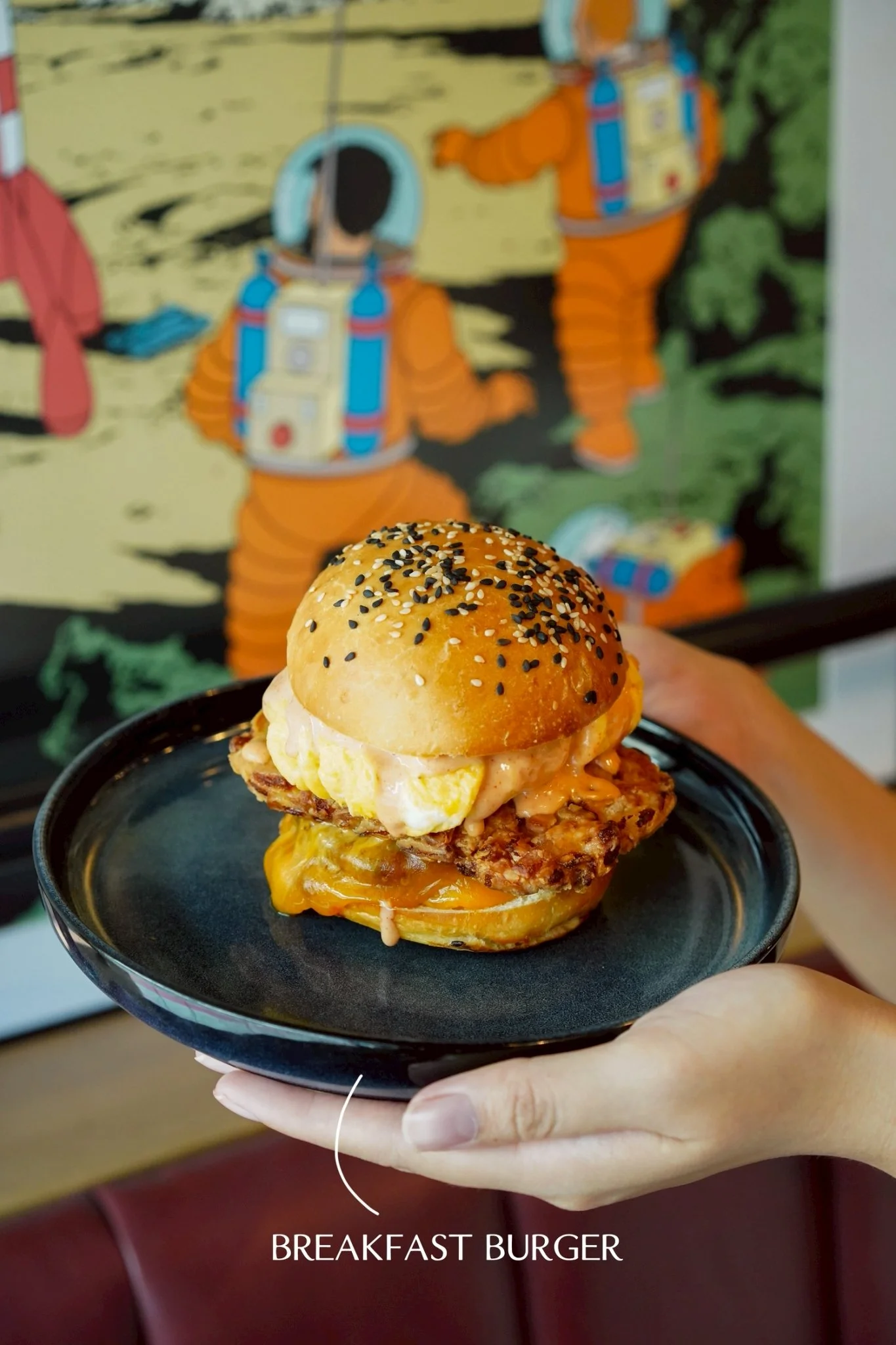 A breakfast burger with a toasted bun topped with black and white sesame seeds, containing scrambled eggs, a fried chicken patty, melted cheese, and sauce, served on a black plate.