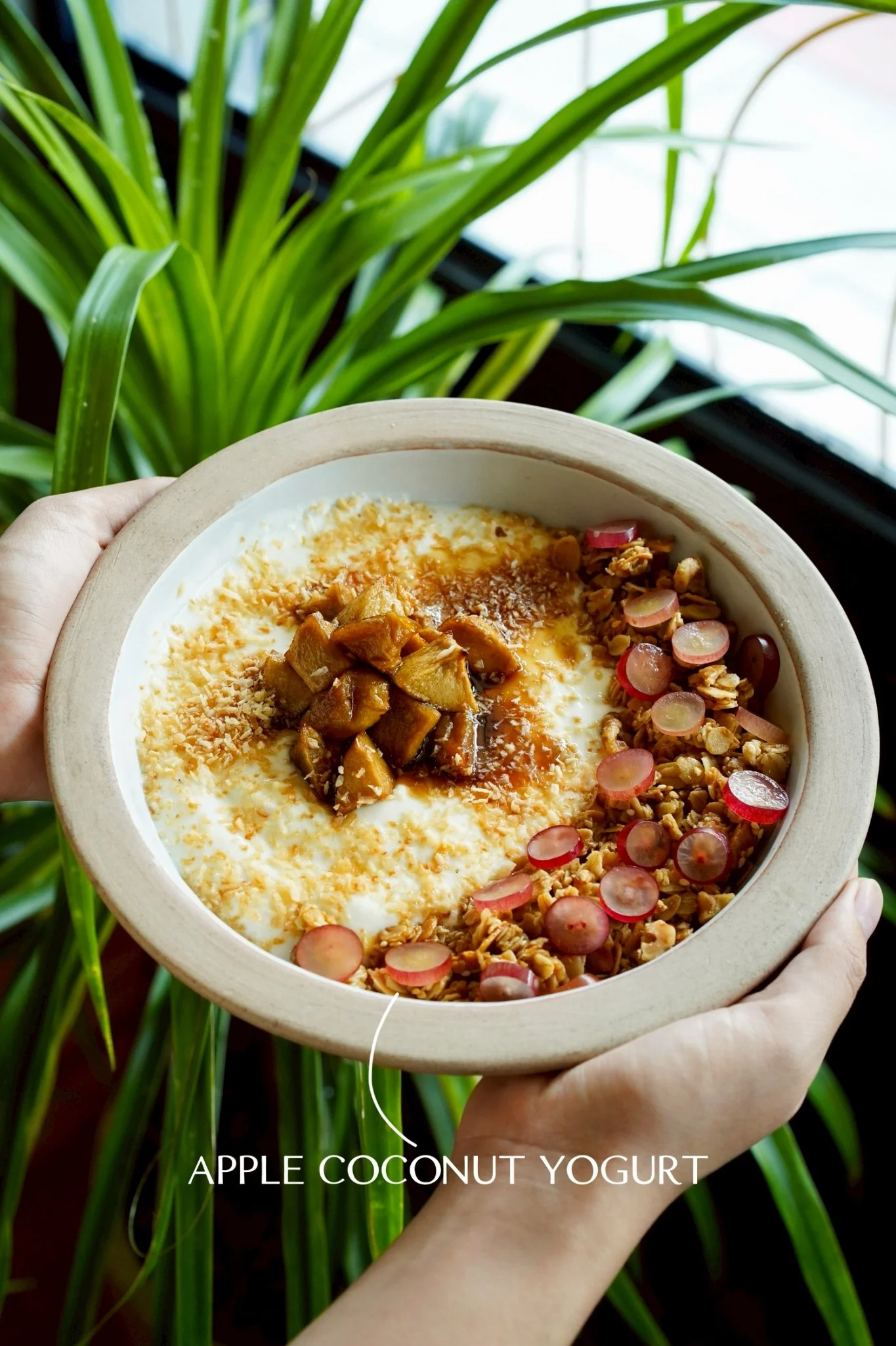 A bowl of apple coconut yogurt topped with granola, sliced radishes, and caramelized apples, held in front of green leafy plants.