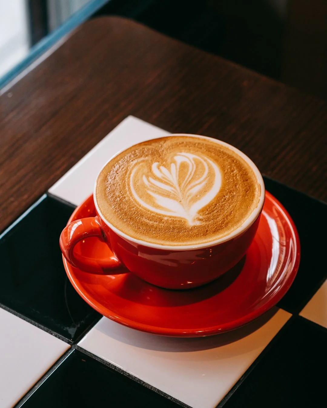 A cup of latte with latte art in a red cup and saucer on a tiled surface