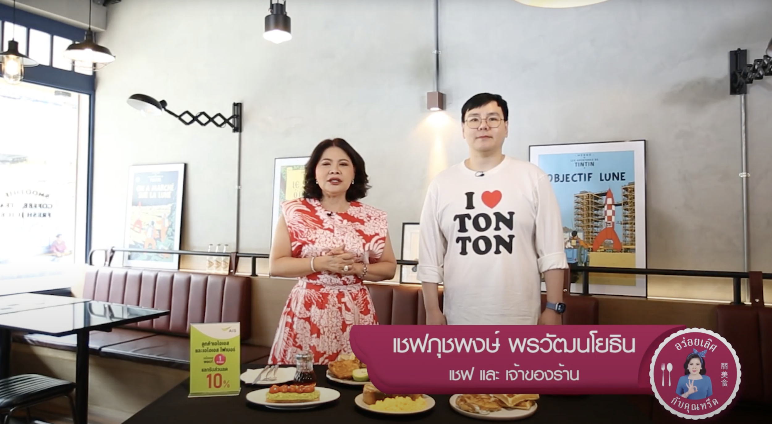 Two people standing behind a table with food in a cafe or restaurant, with framed posters on the wall and a bright window to the left.