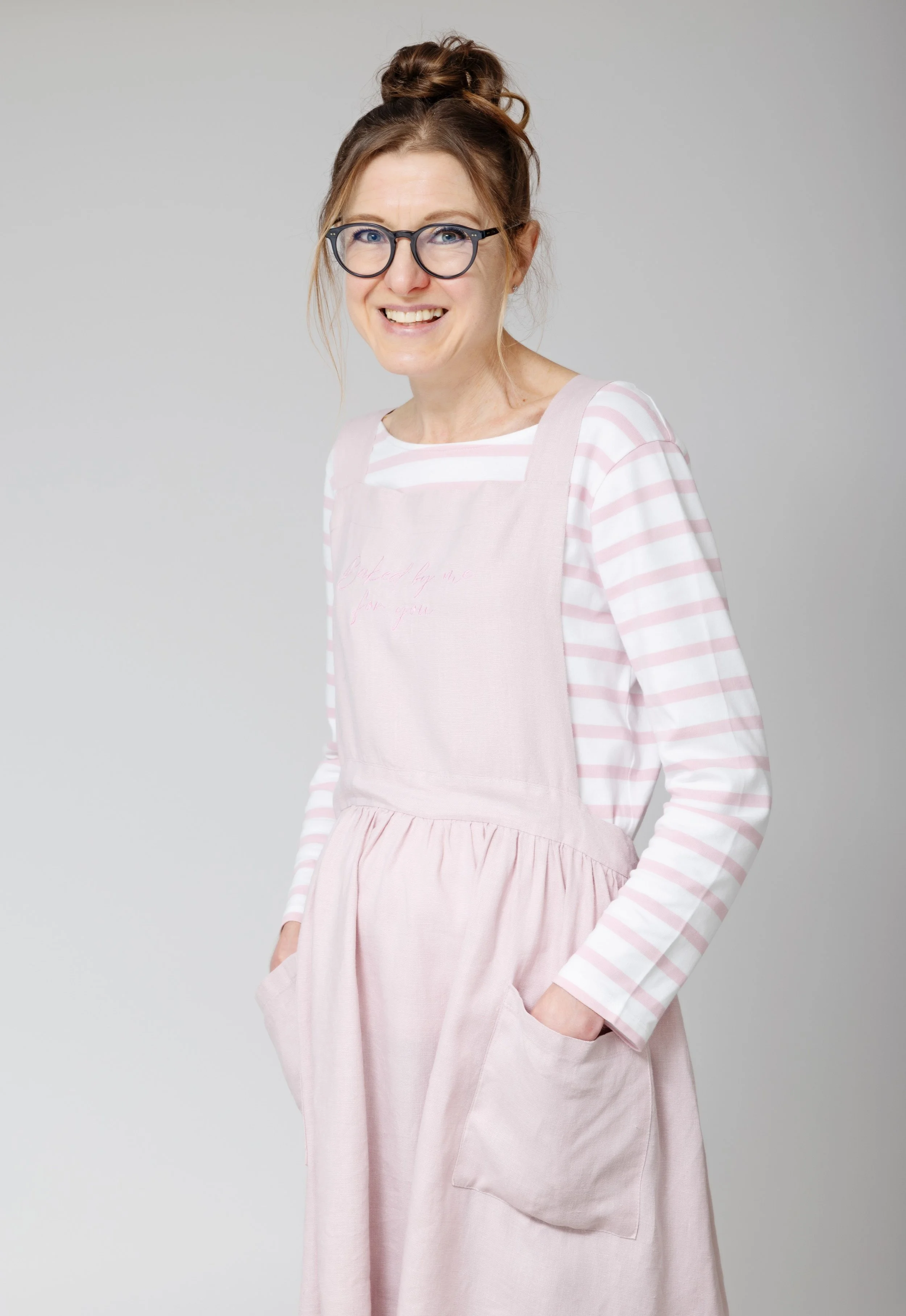 A woman with glasses and a top knot hairstyle wearing a pink apron over a pink and white striped top, smiling at the camera against a plain gray background.