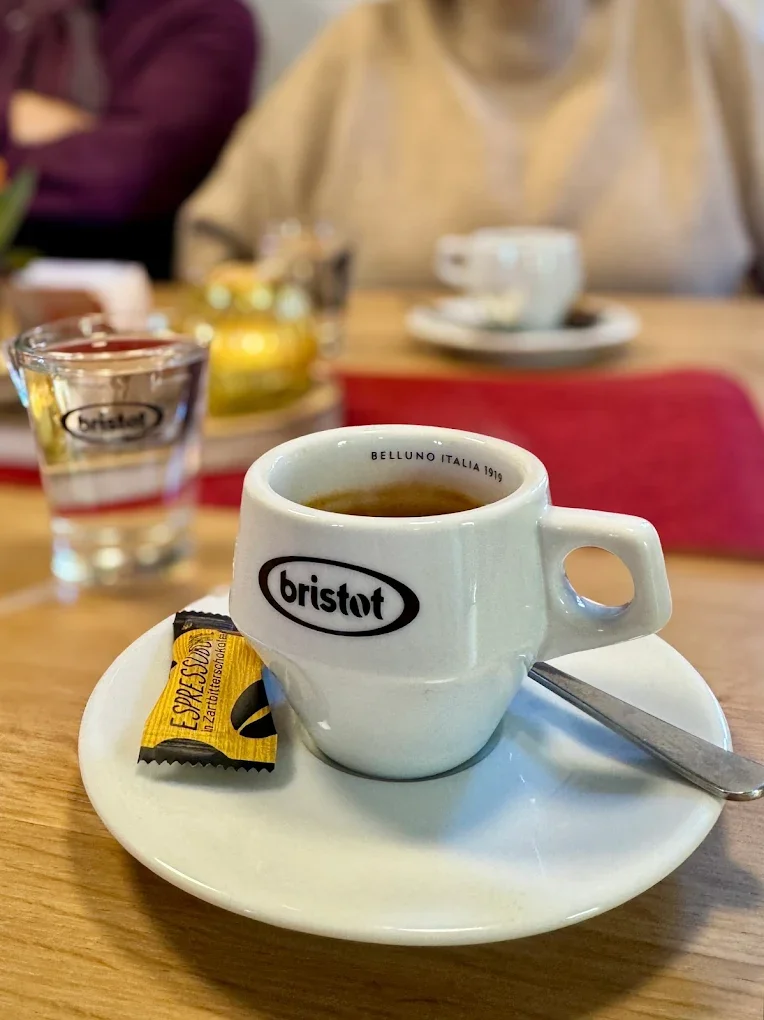 Kaffeetasse mit Kaffee auf einem weißen Teller, darunter eine kleine Speisehilfe, im Hintergrund Gläser und Tassen, auf einem Tisch in einem Café oder Restaurant.