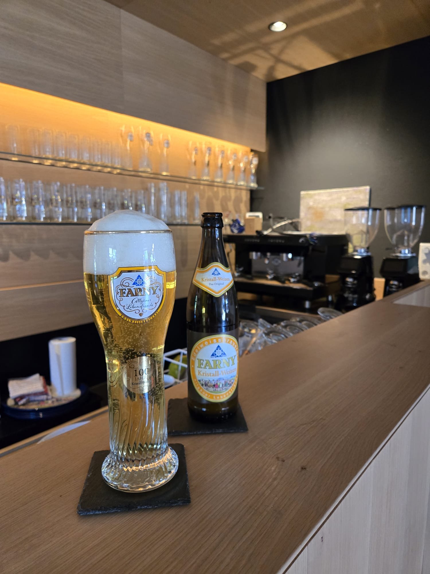 Ein Bierglas und eine Bierflasche auf einer Theke in einer Bar, im Hintergrund Gläser und Bierzapfanlage.