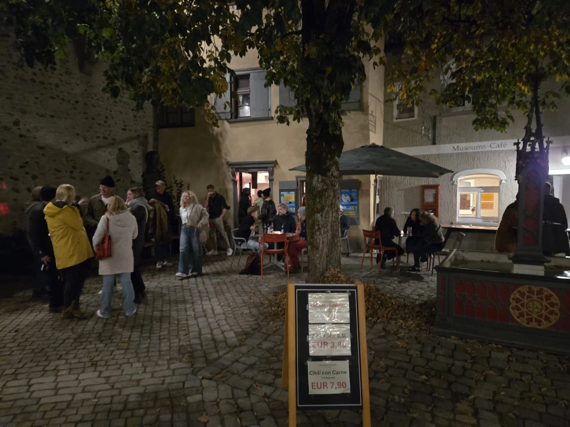 Abendliche Straßenszene mit Menschen, die um einen Tisch im Freien sitzen, und einem Speisekartenständer, der Abendmenüs für EUR 7,90 und EUR 3,80 anbietet. Es gibt Bäume und Gebäude im Hintergrund.