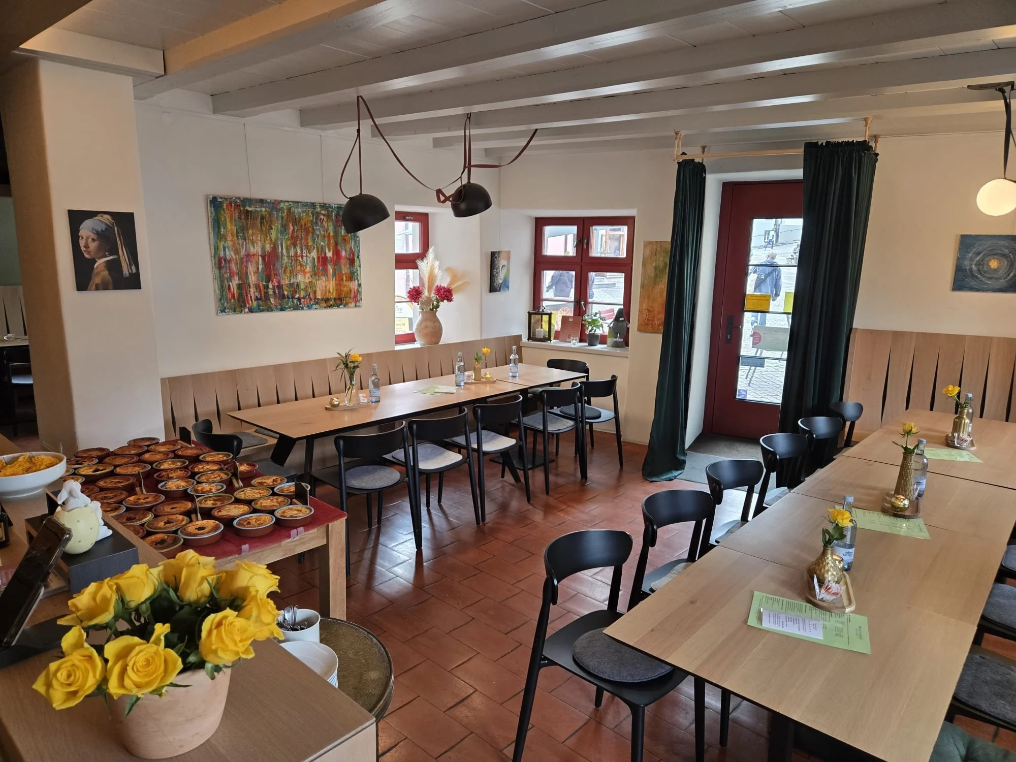 Ein hoher Raum mit Holzbalkendecke, mehreren Tischen und Stühlen, Wanddekorationen, großen Fenstern mit roten Rahmen, grünen Vorhängen und Sonnenlicht, sowie Blumen und Getränke auf den Tischen.