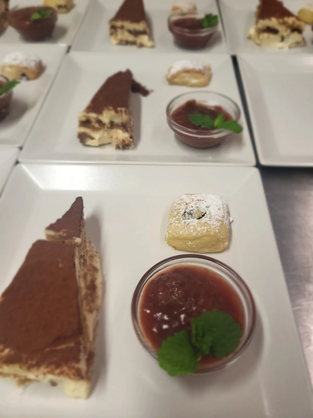 Verschiedene Desserts auf weißen Tellern, inklusive Tiramisu, Zitronenkuchen und Fruchtsauce mit Minzblättern.