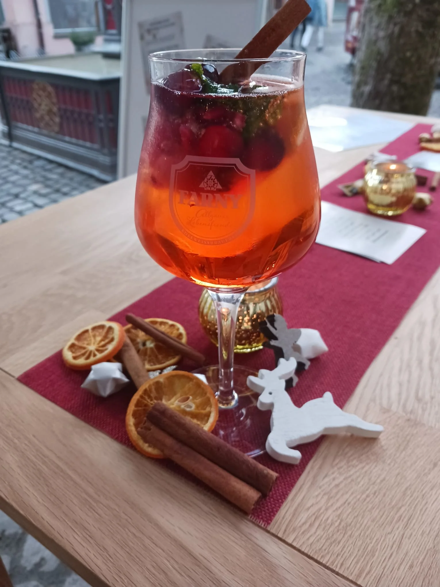Ein Glas mit roter Getränkemischung, Garnitur aus Beeren, Zimtstange und Minze, auf einem Holztisch, umgeben von trockenen Orangenscheiben, Zimtstangen und weihnachtlicher Dekoration.