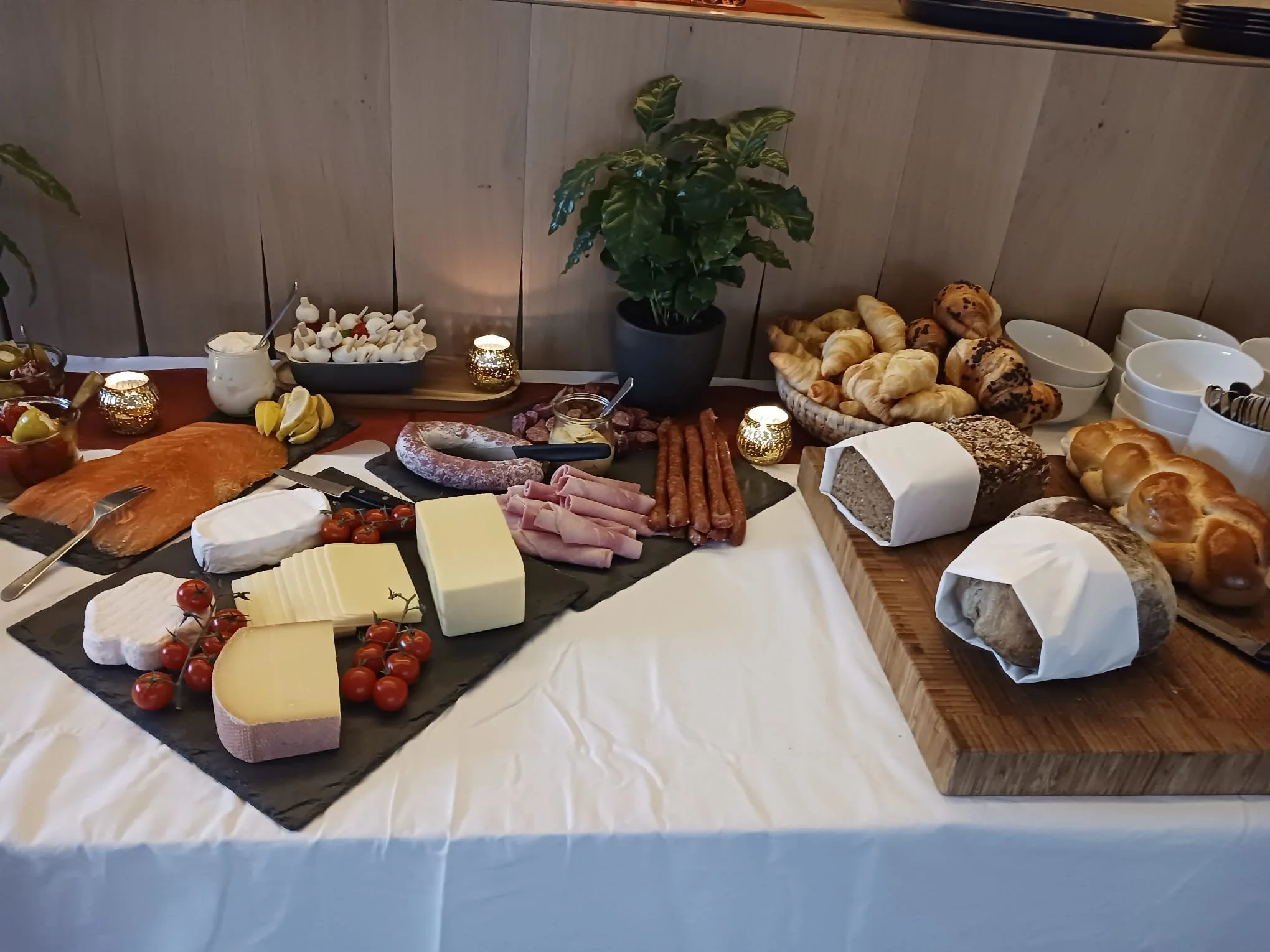 Ein Frühstücksbuffet mit verschiedenen Brotsorten, Käse, Wurst, Tomaten und Obst, dekoriert mit Kerzen und grüner Pflanze, auf einem Tisch mit weißer Tischdecke.