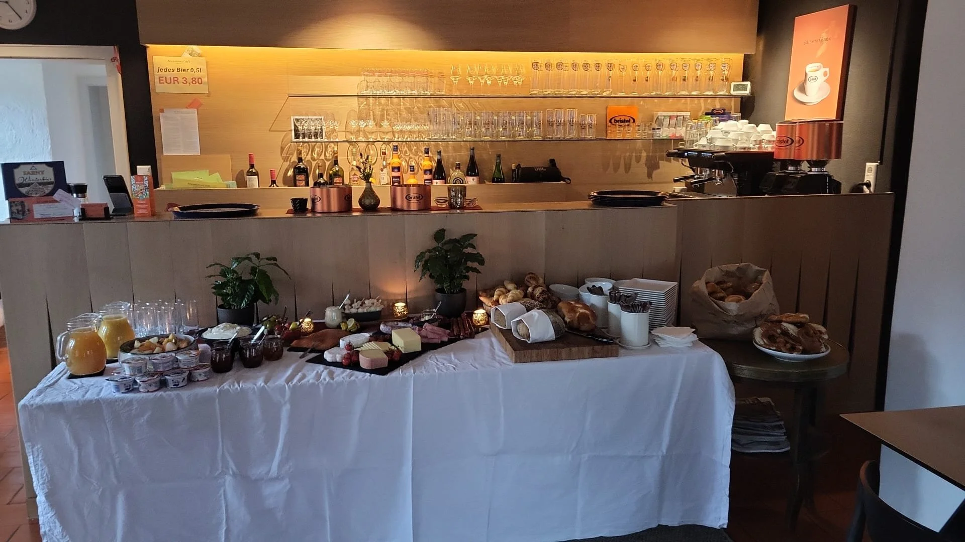 Frühstücksbuffet mit verschiedenen Getränken, Käse, Wurst, Gebäck, Joghurt, Marmelade, und Kaffee in einem Café oder Hotel. Hintergrund mit Gläsern und Flaschen auf Holzregal.