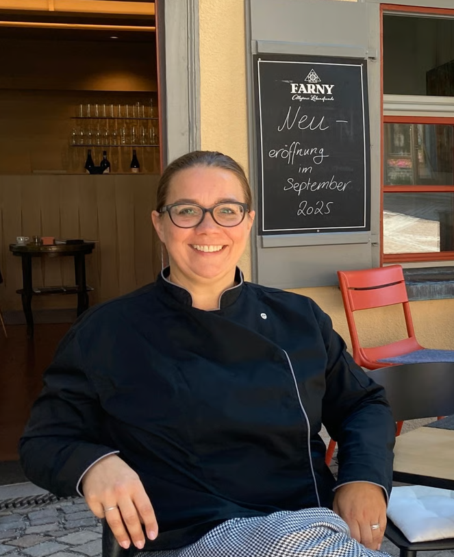 Eine lachende Frau in einer schwarzen Kochjacke sitzt draußen vor einem Restaurant, daneben ein schwarzes Schild mit weißen Schriftzügen, das auf eine Neueröffnung im September 2025 hinweist.