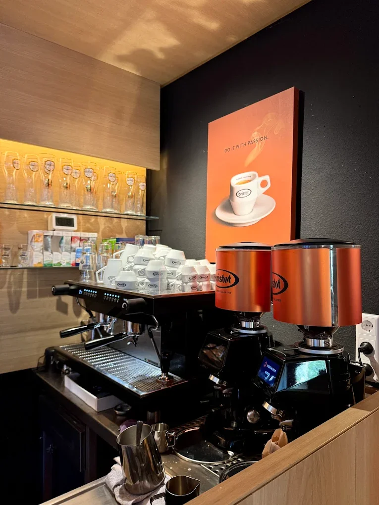 Eine Kaffee-Bar mit Espressomaschinen, weißen Kaffeetassen, ein Display, Gläser auf einer obersten Regalblatte und einem Poster mit einer dampfenden Kaffeetasse auf schwarzer Wand.