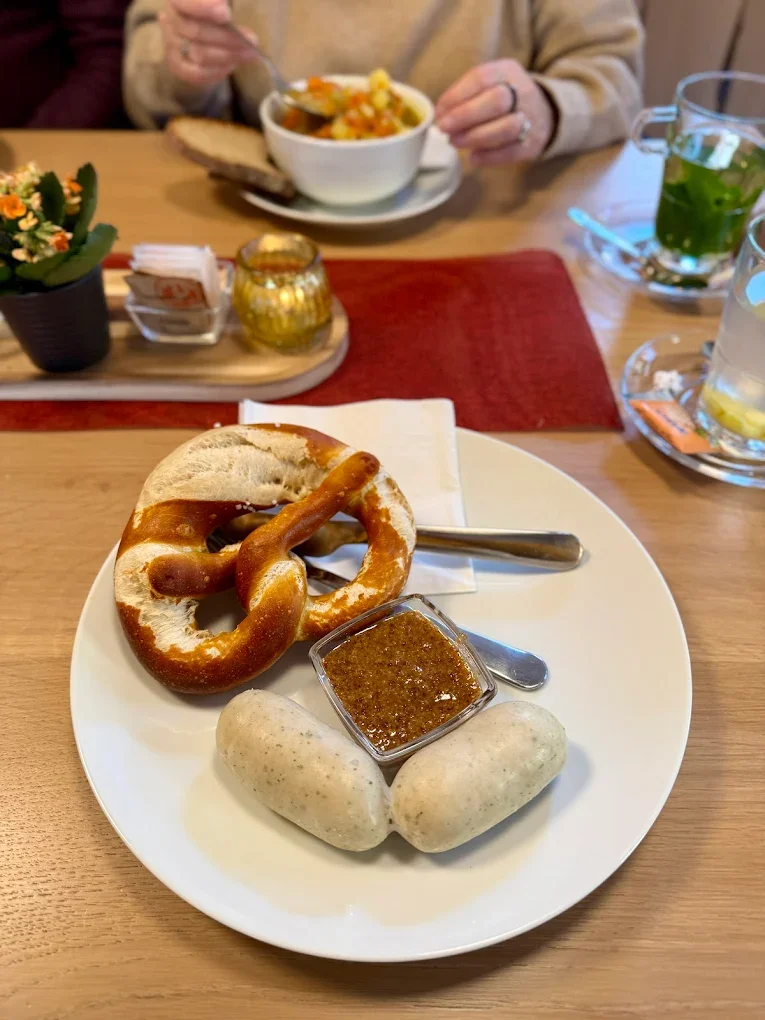Deutsch-frühstück mit Brezeln, Weißwürsten und süßem Senf auf einem weißen Teller, im Hintergrund eine Person mit Suppe und Getränke auf einem Holztisch.