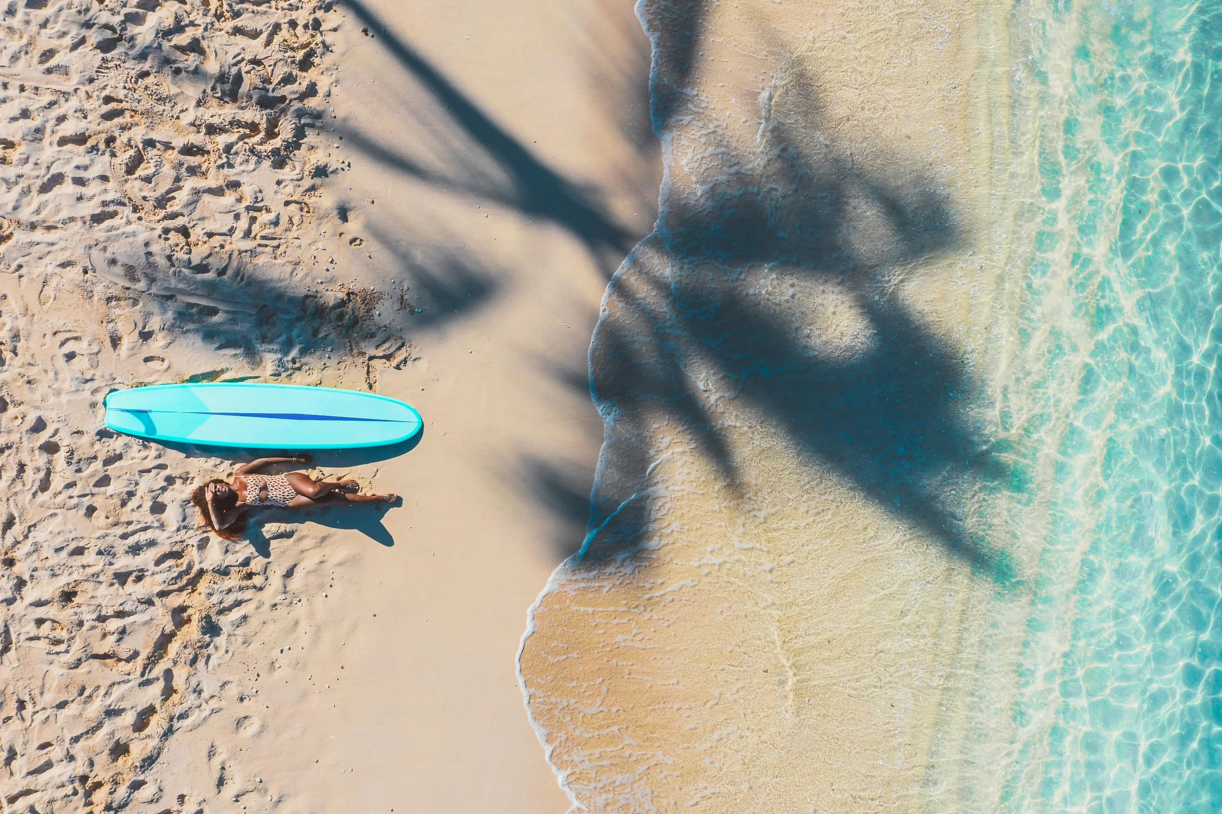 Une femme allongée sur la plage près d'une planche de surf turquoise, avec des palmiers projetant des ombres sur le sable, à proximité de l'eau claire de la mer.