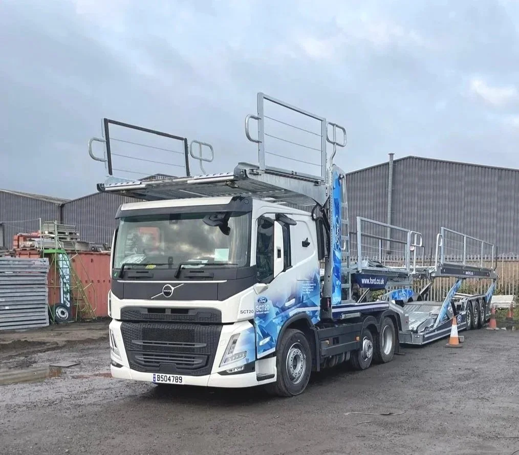 A large white truck with blue graphics, designed for transporting vehicles, is parked on a dirt lot near industrial buildings, with orange traffic cones nearby.