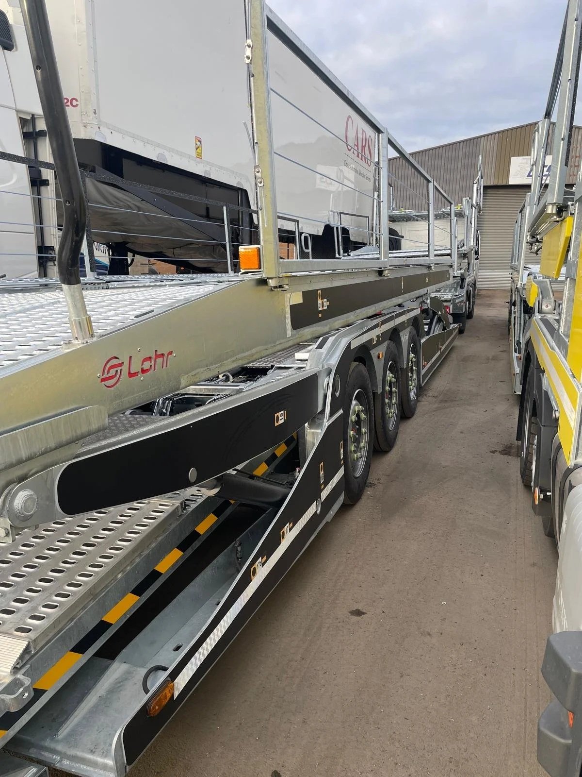 A flatbed truck with a low-loader trailer, parked in a yard alongside other trucks, with a warehouse or industrial building in the background.