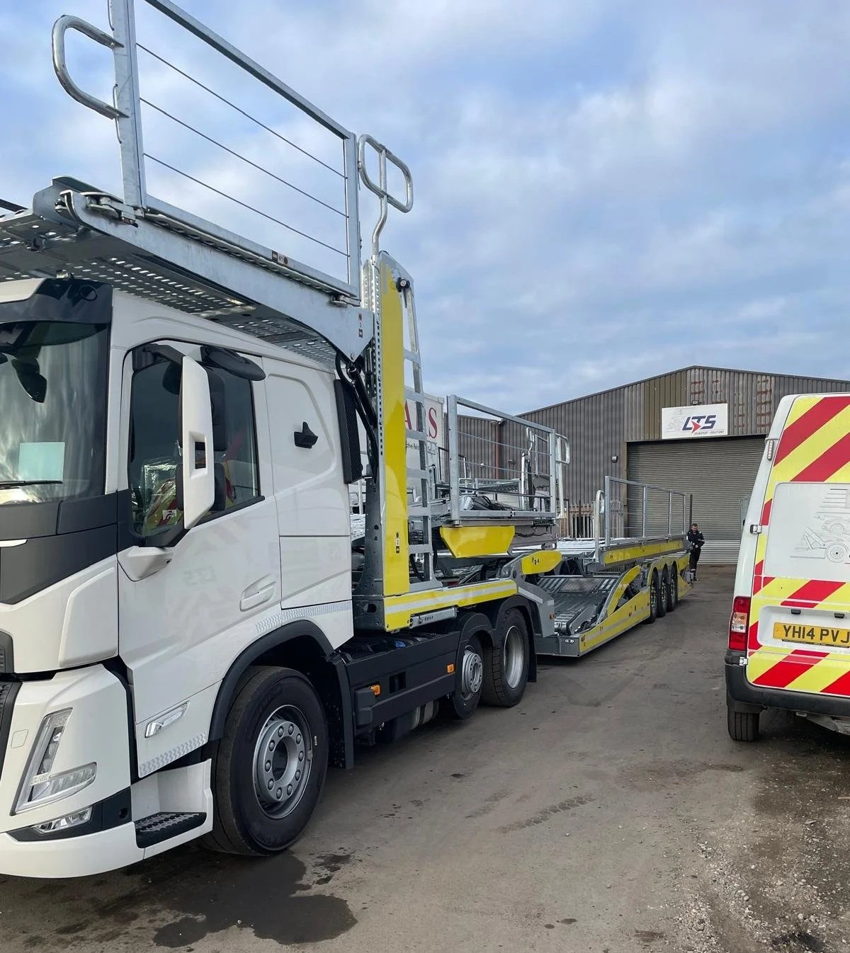 A large white truck with a yellow and gray loading platform and a metal safety railing, parked outside a warehouse with a closed roll-up door. There is a white van with red and yellow reflective stripes nearby and a person standing near the warehouse