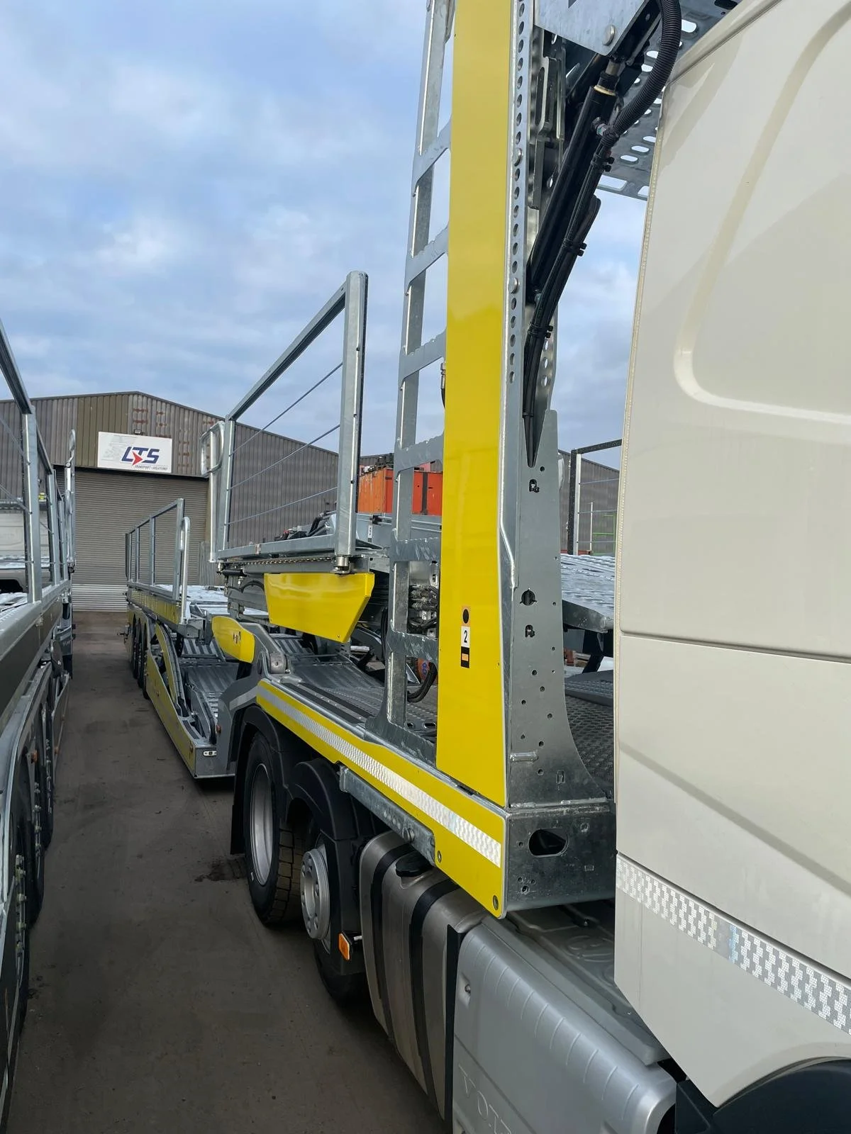 A close-up view of a flatbed truck with a yellow safety barrier, parked outdoors near a warehouse, with other trucks and a cloudy sky visible in the background.