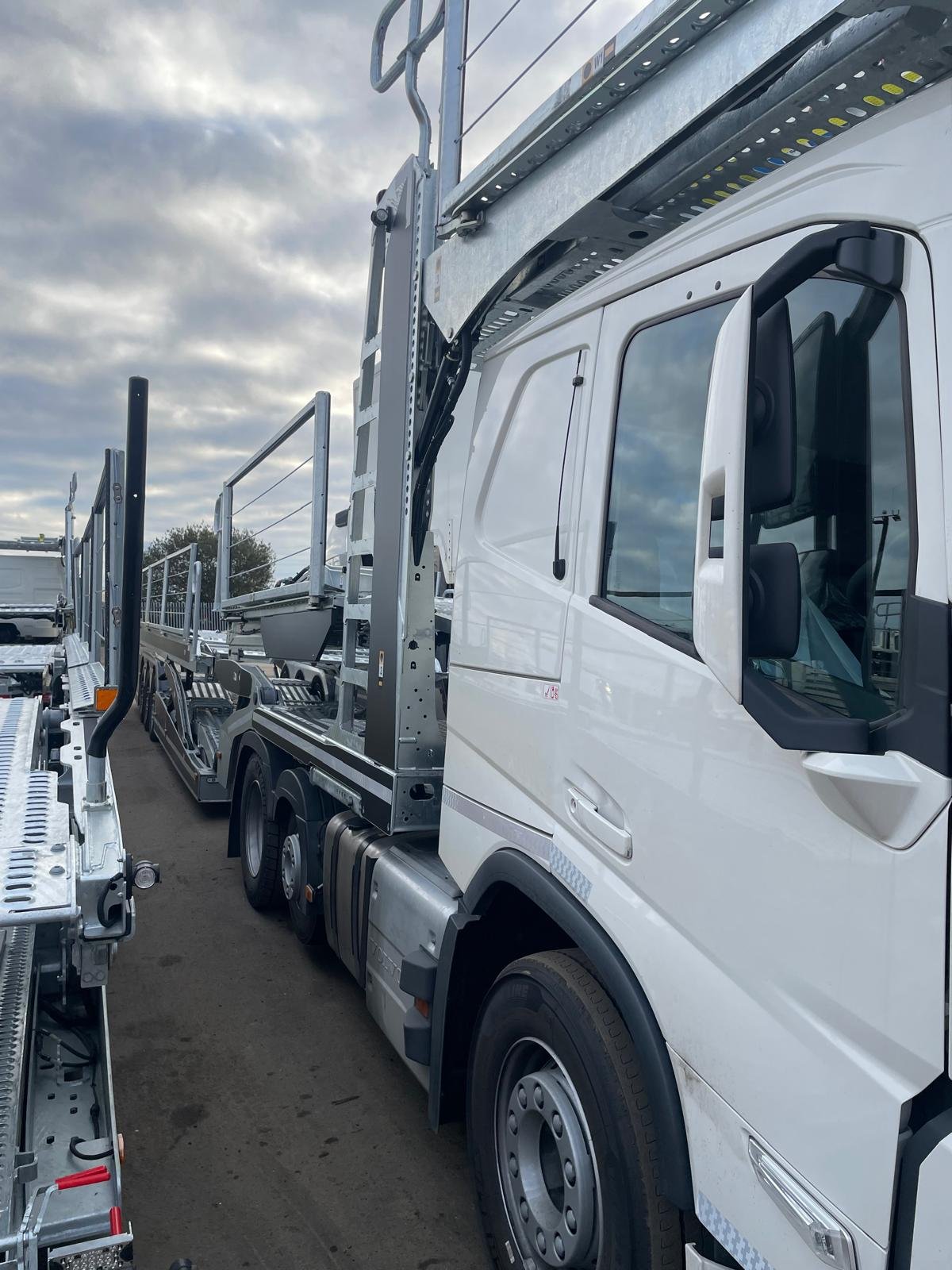Side view of a white car carrier truck parked outdoors on a cloudy day, with similar vehicles and a dirt ground in the background.