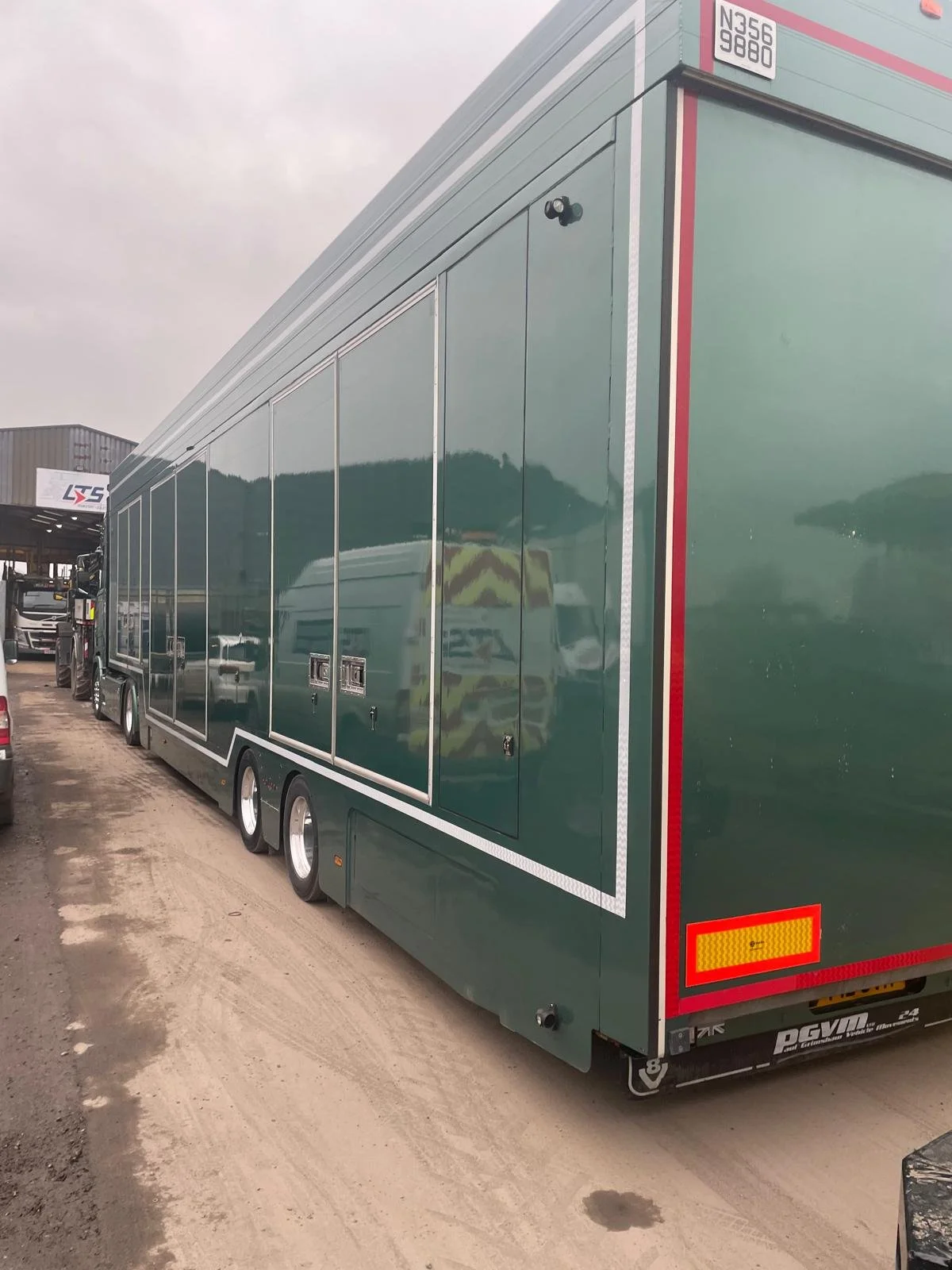 A large green truck parked on a dirt lot, with a reflective surface showing the environment and a reflection of a person wearing a yellow safety vest.