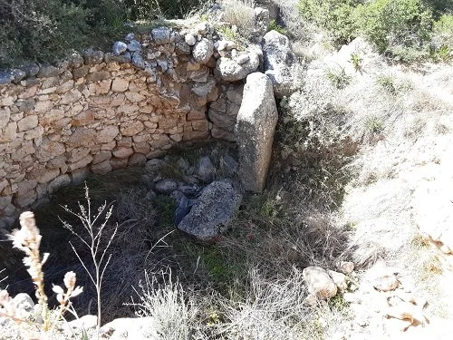 A small rocky stone structure with a curved wall, situated in a dry, brushy area with sparse vegetation and large rocks nearby.