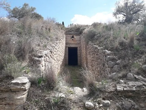 Underground tunnel with stone walls and a small opening, surrounded by dry grass and shrubs, in a natural landscape under a cloudy sky.