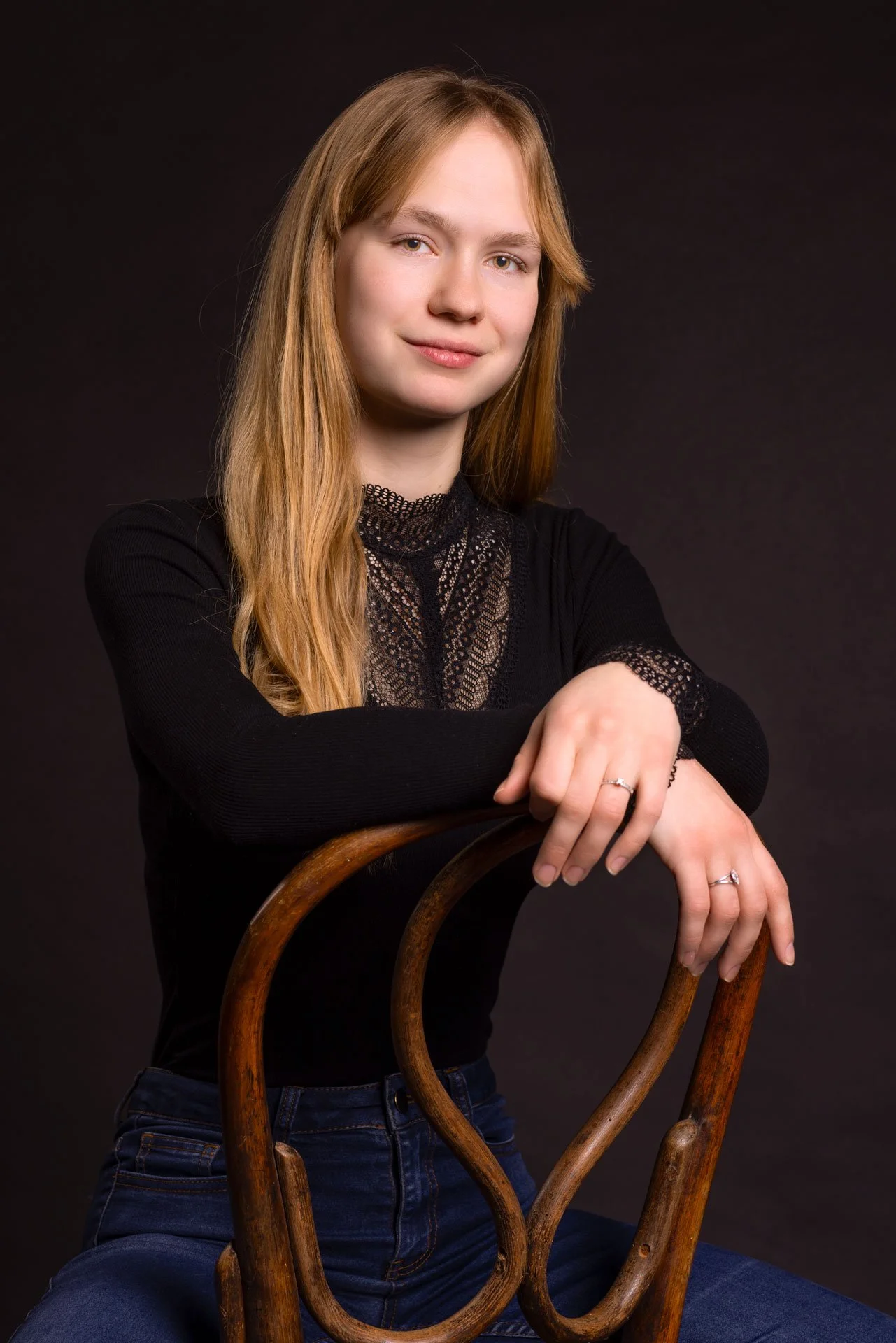 Portræt af en ung kvinde med langt, blond hår, iført en sort top med blonde detaljer, siddende på en træstol mod en sort baggrund.