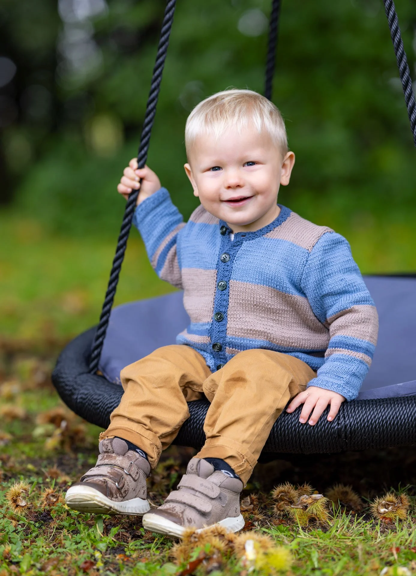 En glad dreng med blond hår sidder på en gynge udendørs i naturen, omgivet af grønt græs og træer.