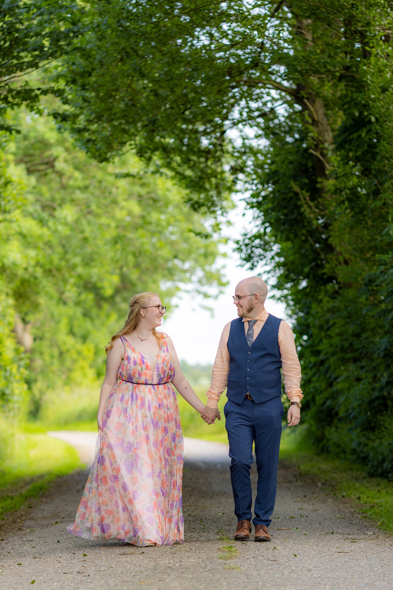 En glad par går hånd i hånd på en sti omgivet af grønne træer.