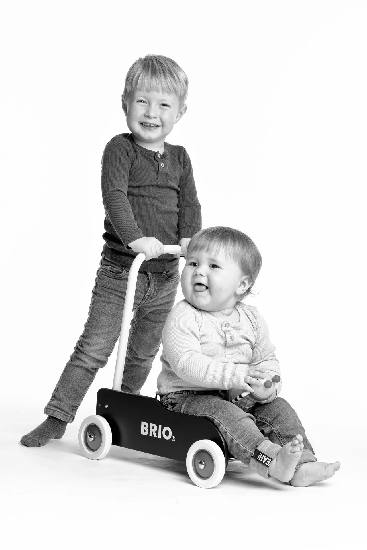 To unge børn, et sidder i en legetøjs-stol og holder en legetøjsfigur, den anden står bag med en walker, begge smiler.