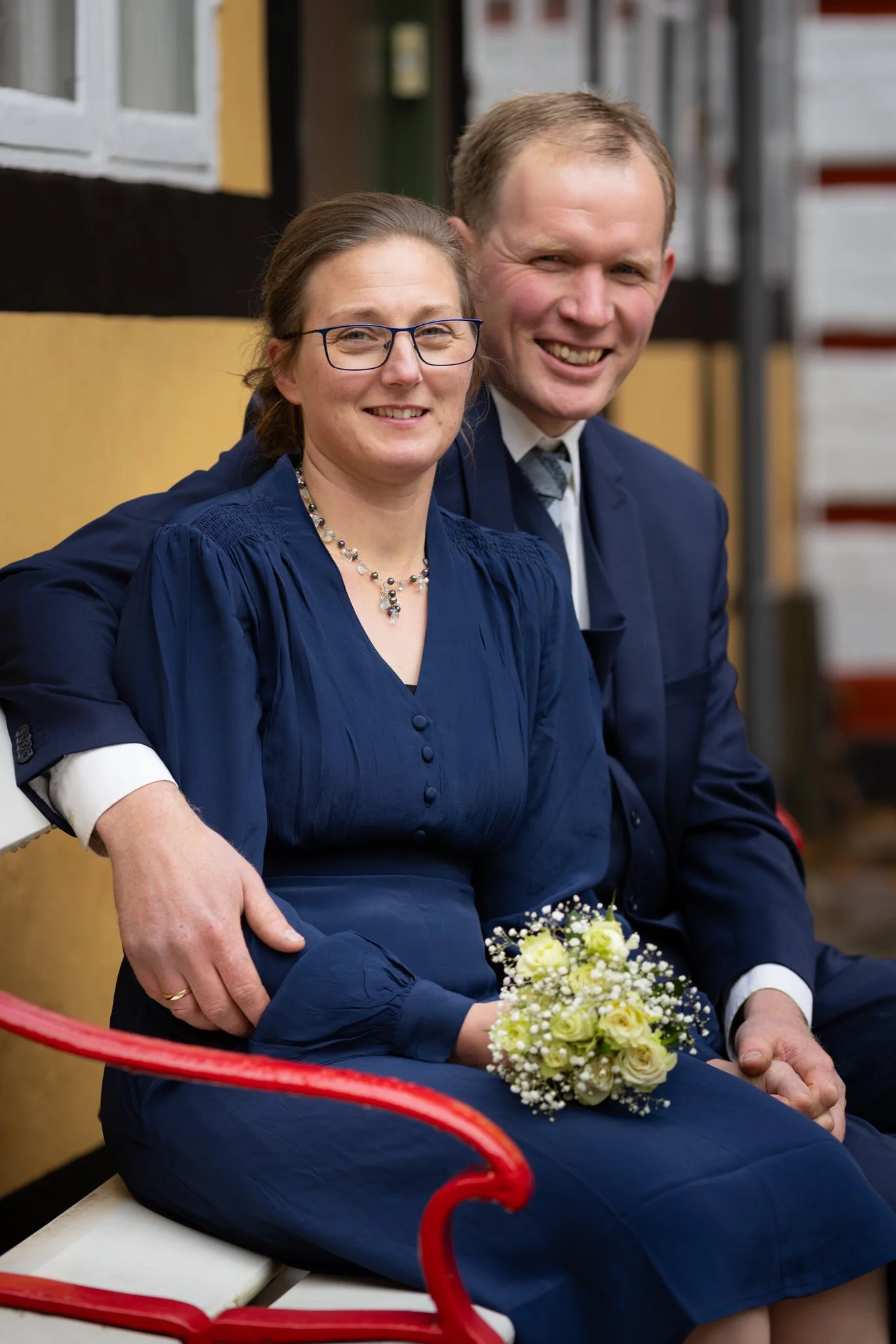 Et smilende par sidder på en bænk udenfor, den kvinde holder en lille buket af hvide blomster, begge er iført blå tøj, manden har en skjorte og slips.