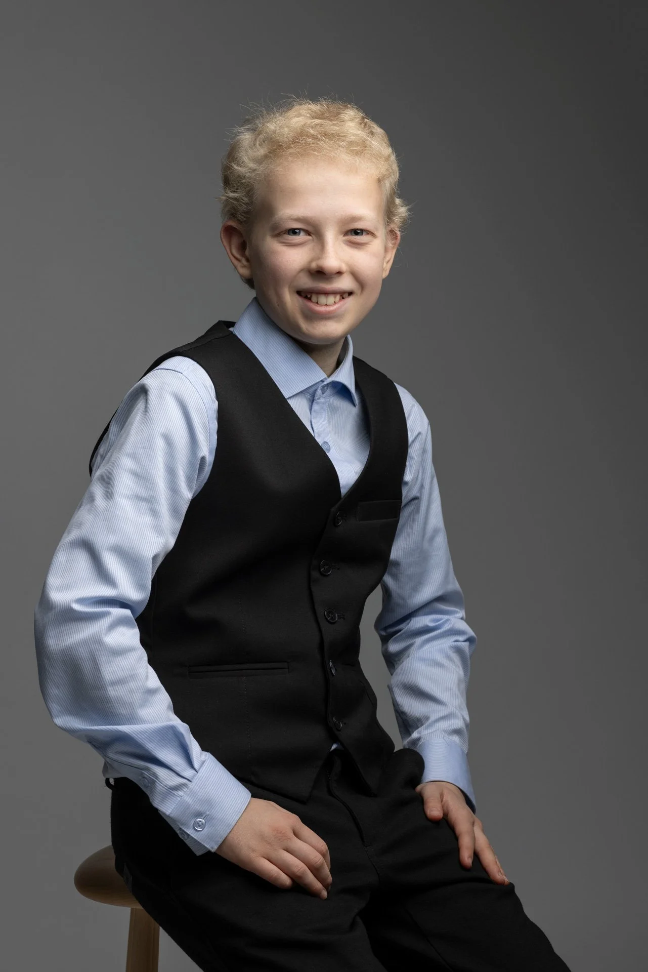 Ung mand sidder på en træstol foran en grå baggrund, iført blå skjorte og sort vest.