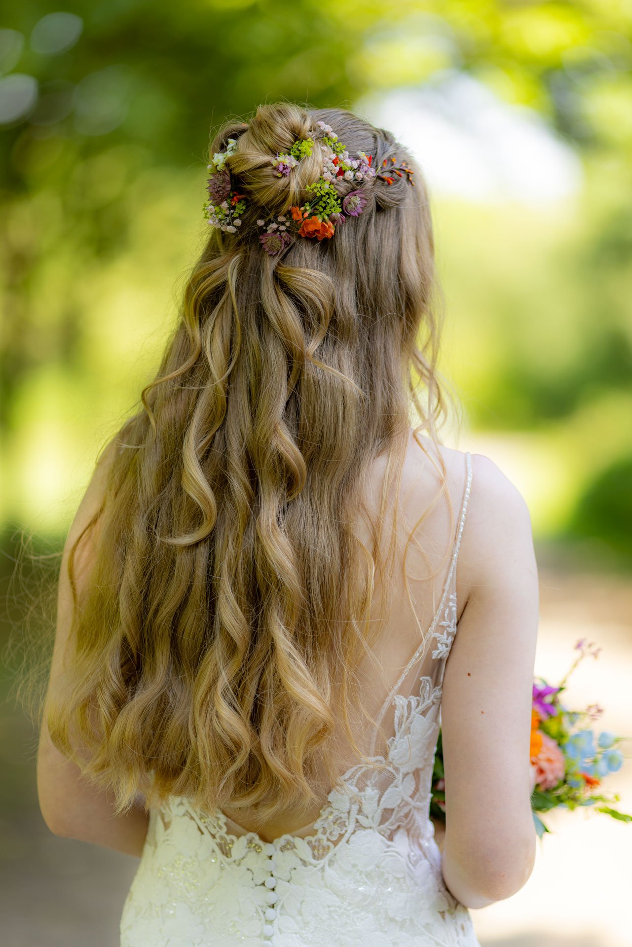 En brud med lang, krøllet, blond hår, båret i en halv opstillet med blomsterdekoration, set udefra i et lyst, grønt udendørs miljø.