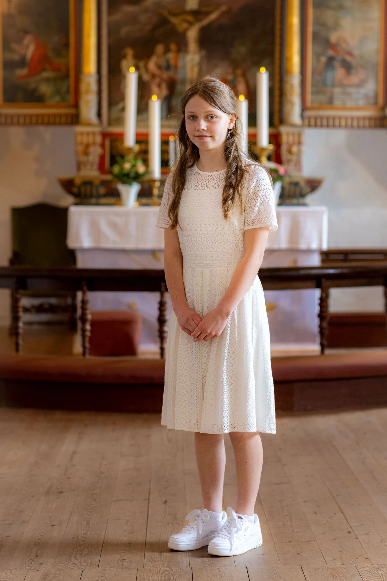 En ung pige i en hvid kjole og sneakers står inde i en kirke foran alteret med lys og religiøse billeder i baggrunden.