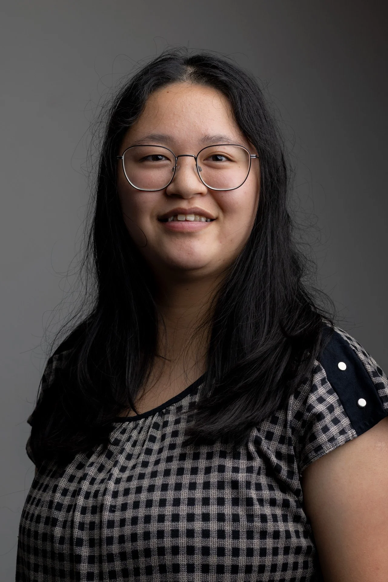 Portræt af en ung kvinde med briller og mørkt, langt hår, iført en sort og beige checket T-shirt med sorte detaljer med perler, smilende mod kameraet, med en grå baggrund.