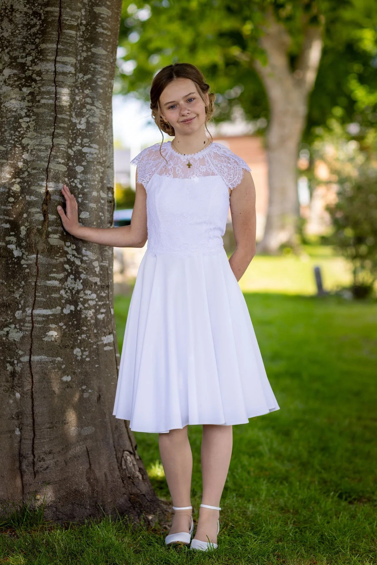 Ung pige i hvid kjole står ved et træ i en park på en solskinsdag.