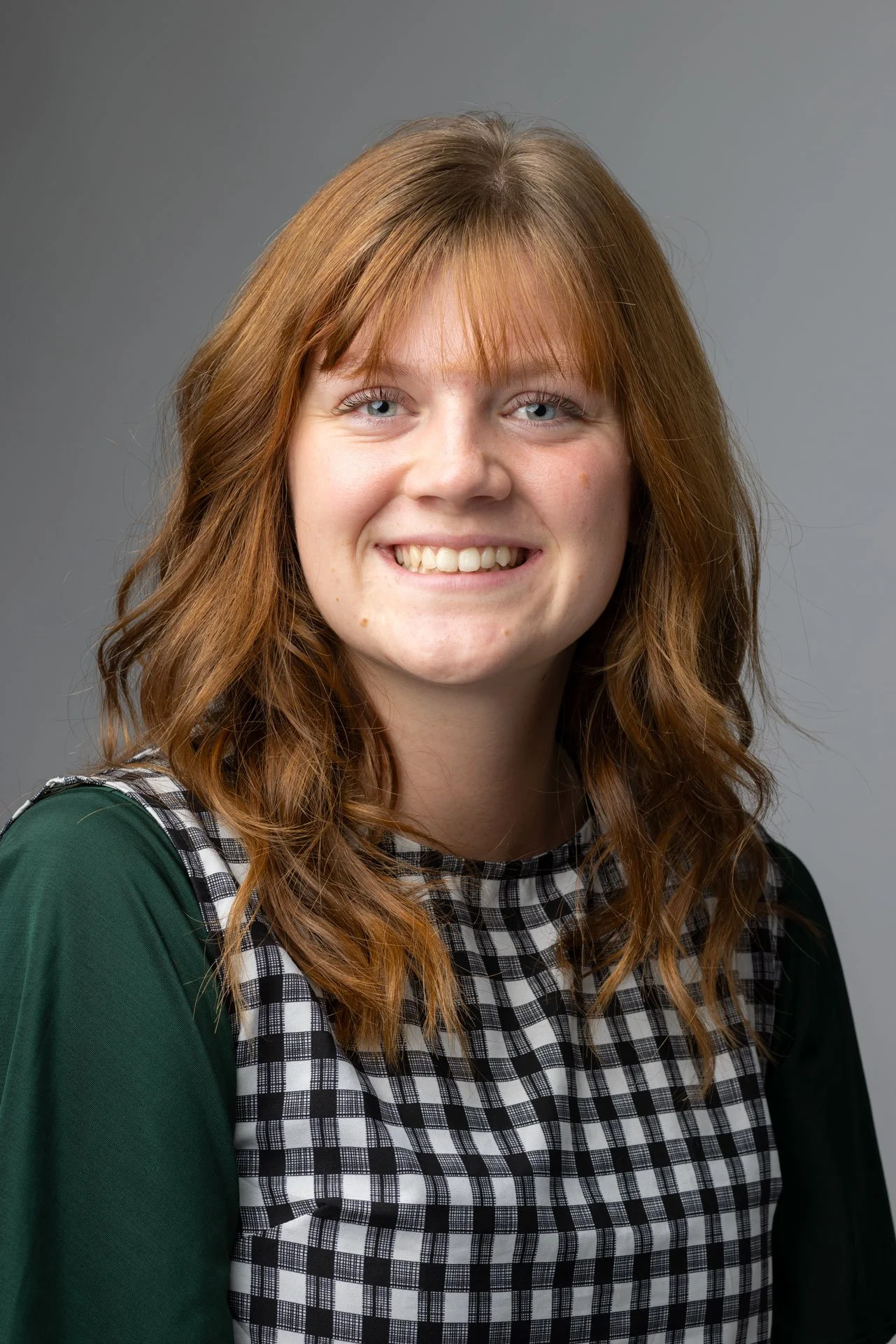 Et portræt af en smilende ung kvinde med lyseblå øjne, bølget, rødbrunt hår, iført en grøn top og sort-hvid ternet skjorte mod en grå baggrund.