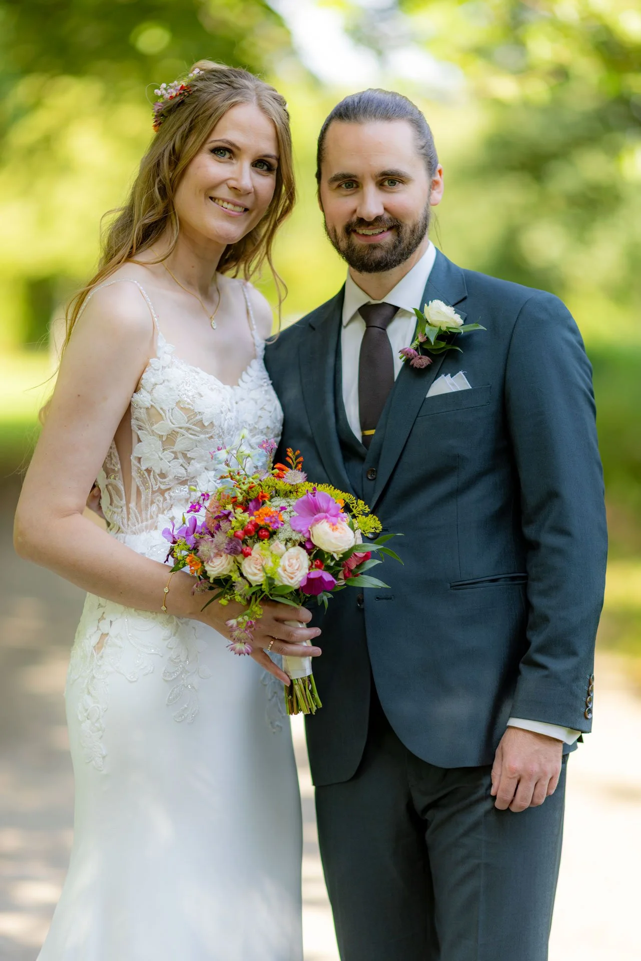 En brud i en hvid brudekjole holder en blomsterbuket, står ved siden af en brudgom i en mørkeblå jakkesæt, udendørs med grønne træer i baggrunden.