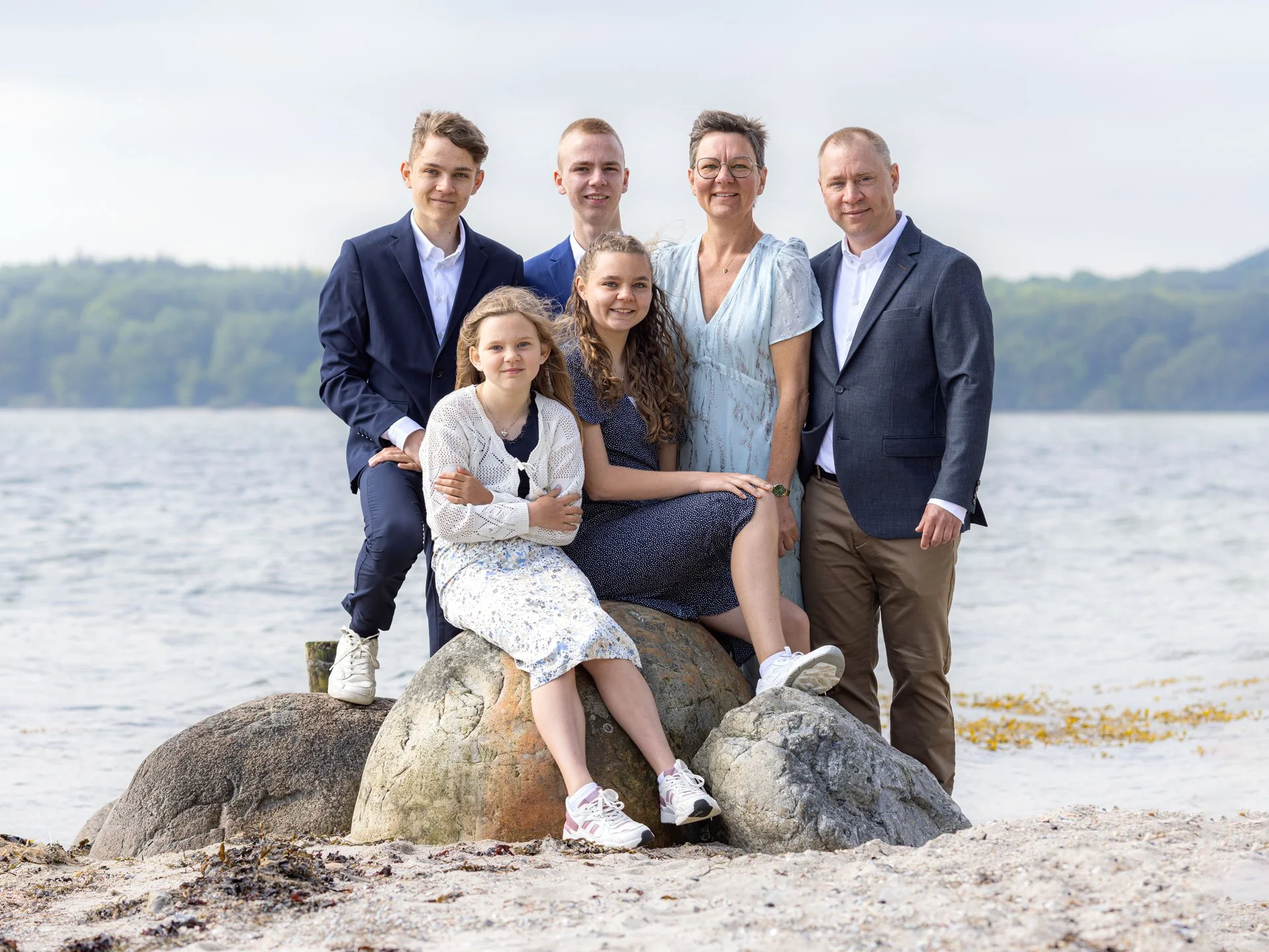 En familie på syv personer står og sidder på store sten ved en strand med vand i baggrunden. Familien består af tre børn, to voksne og to unge voksne, alle smilende og klædt i semi-formelle tøj.