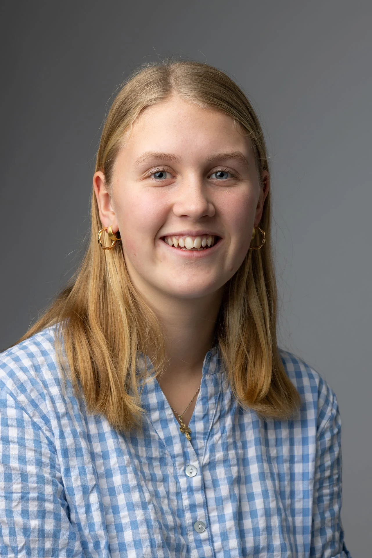 Portræt af en smilende ung pige med blonde hår og blå øjne, iført en blå og hvid ternet skjorte, guld øreringe og et halskæde med en kors-vedhæng, mod en grå baggrund.