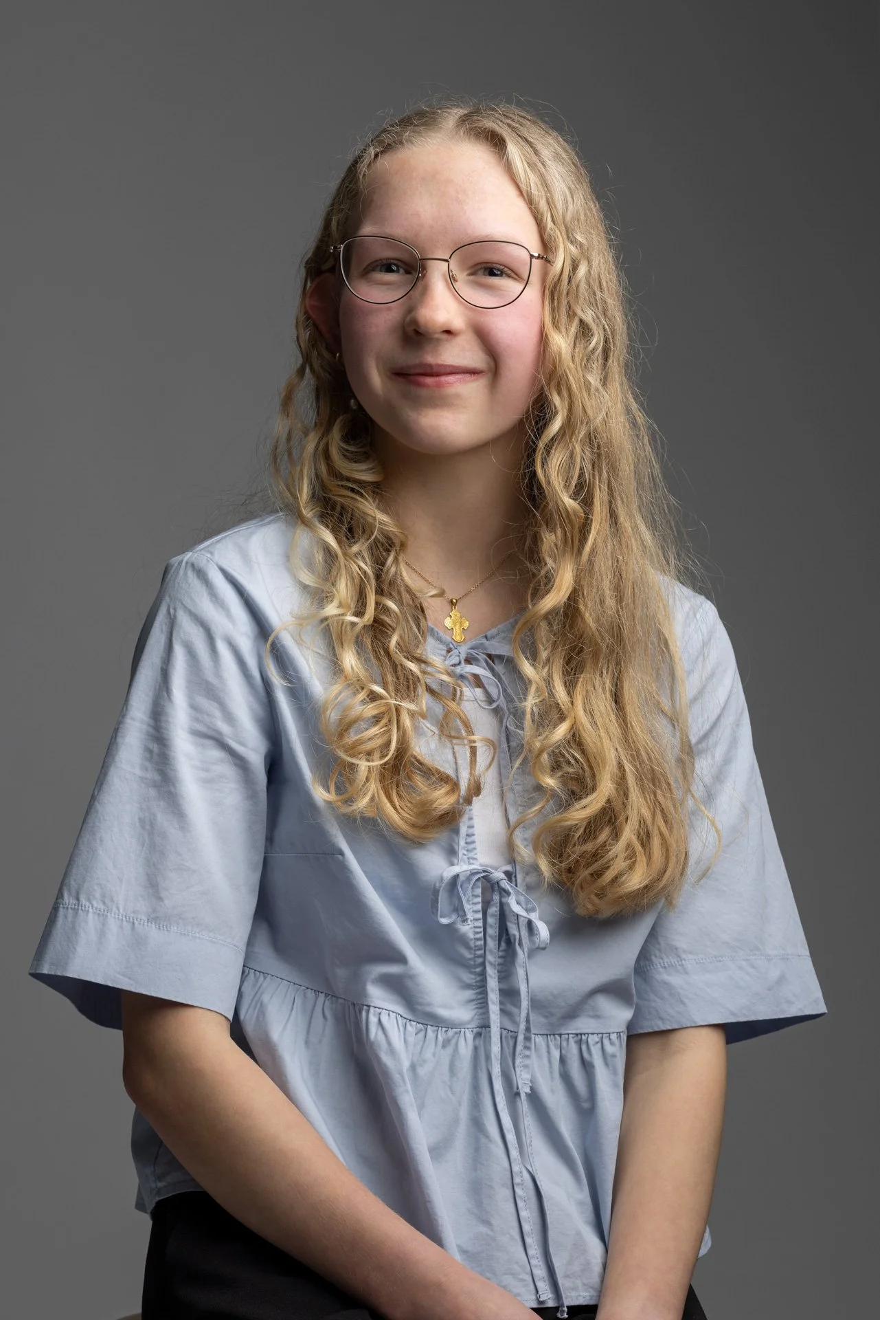 Ung kvinde med blond, krøllet hår, briller og en guldsmykke, smilende mod kamera mod en grå baggrund.