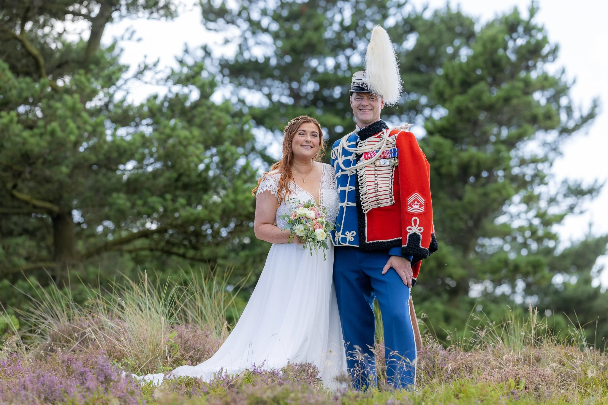 En kvinde i en hvid brudekjole holder en blomsterbuket, sammen med en mand i en uniform med stor hvide fjer på hovedet, begge står på en lavning omgivet af grønne træer og lavendel.