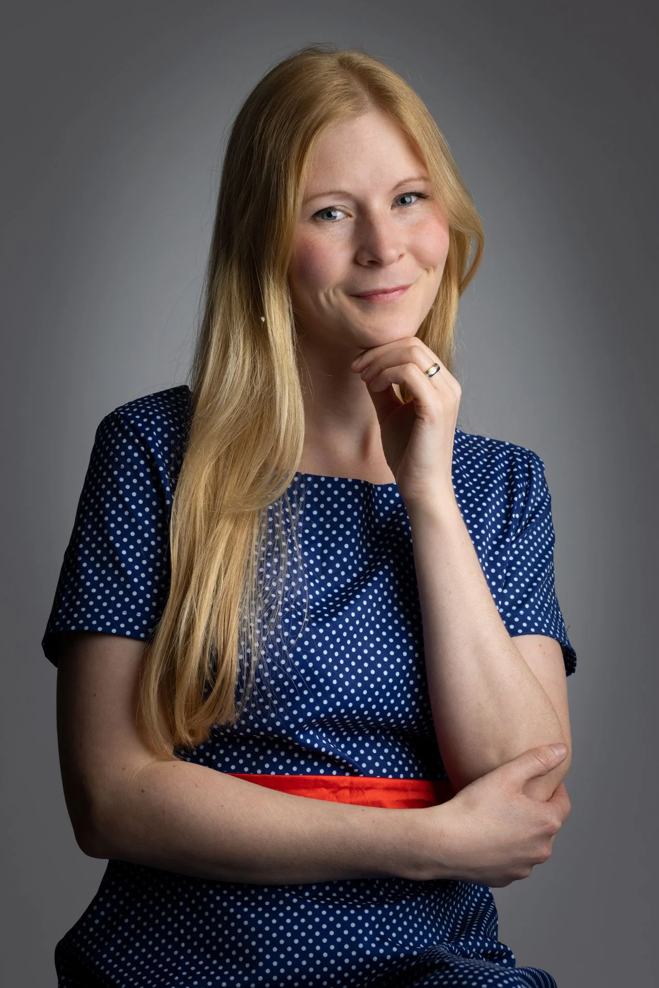 Kvindelig person med lang blond hår, iført blå kjole med hvide polka dots, smiler let, har en finger ved hagen og ser direkte mod kameraet.