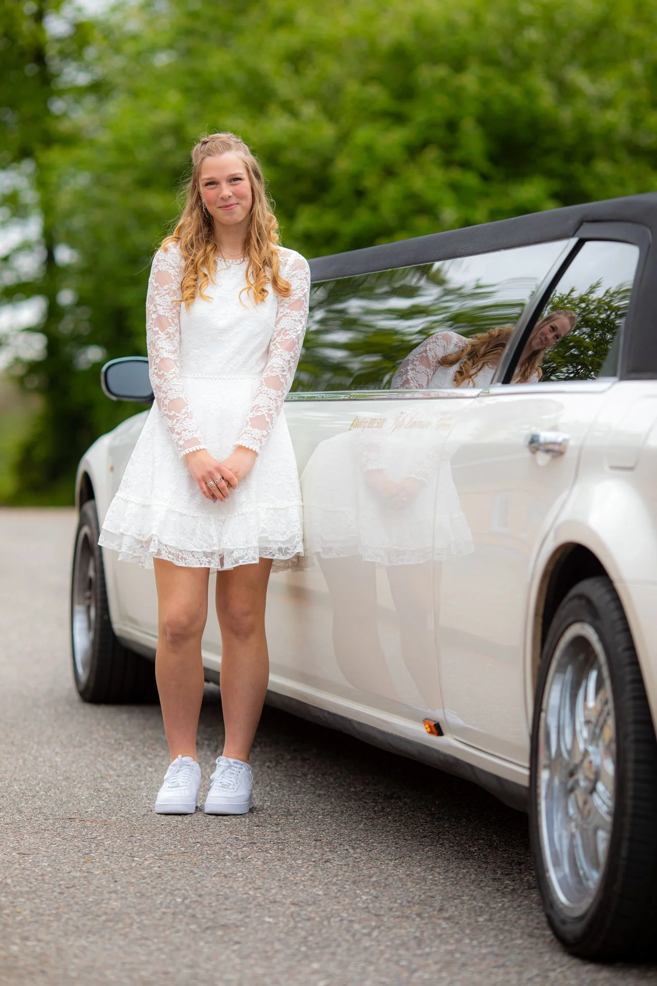 En ung kvinde i hvid brudekjole står ved siden af en hvid cabriolet bil på en vej omgivet af grønne træer.