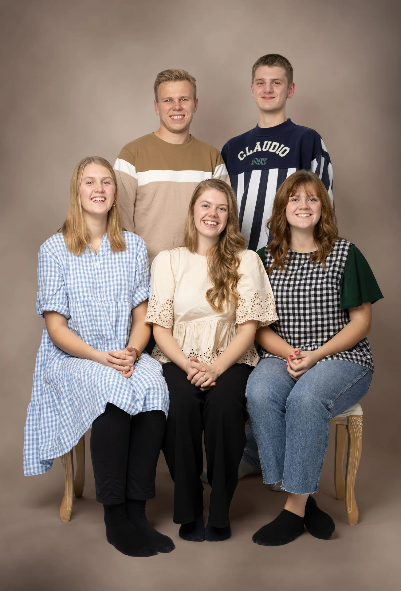 Gruppebillede af fem unge mennesker, tre piger sidder foran, to mænd står bagved, alle smiler mod kameraet, mod en neutral beige baggrund.