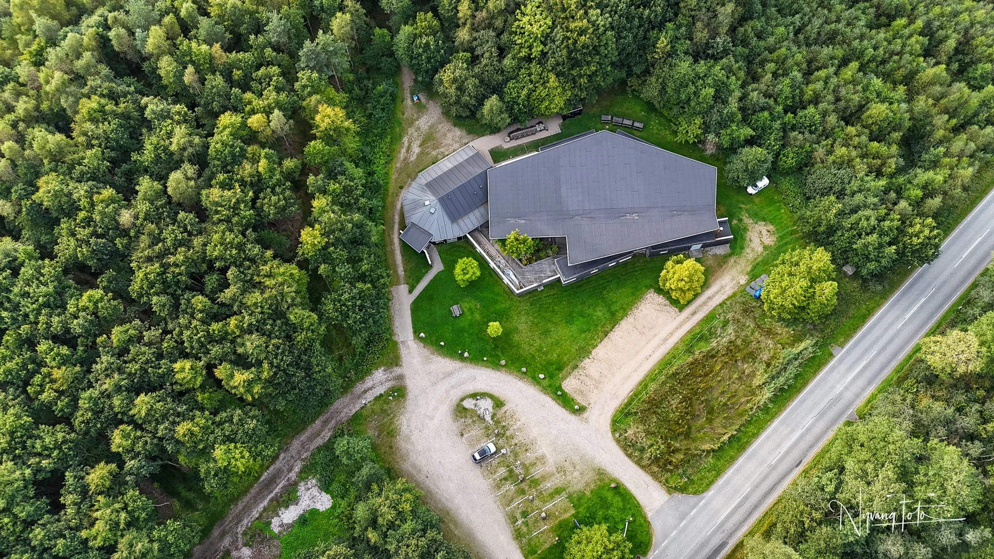 Overhead billede af et moderne hus omgivet af grønne træer, en parkeringsplads med en bil, og en landevej.