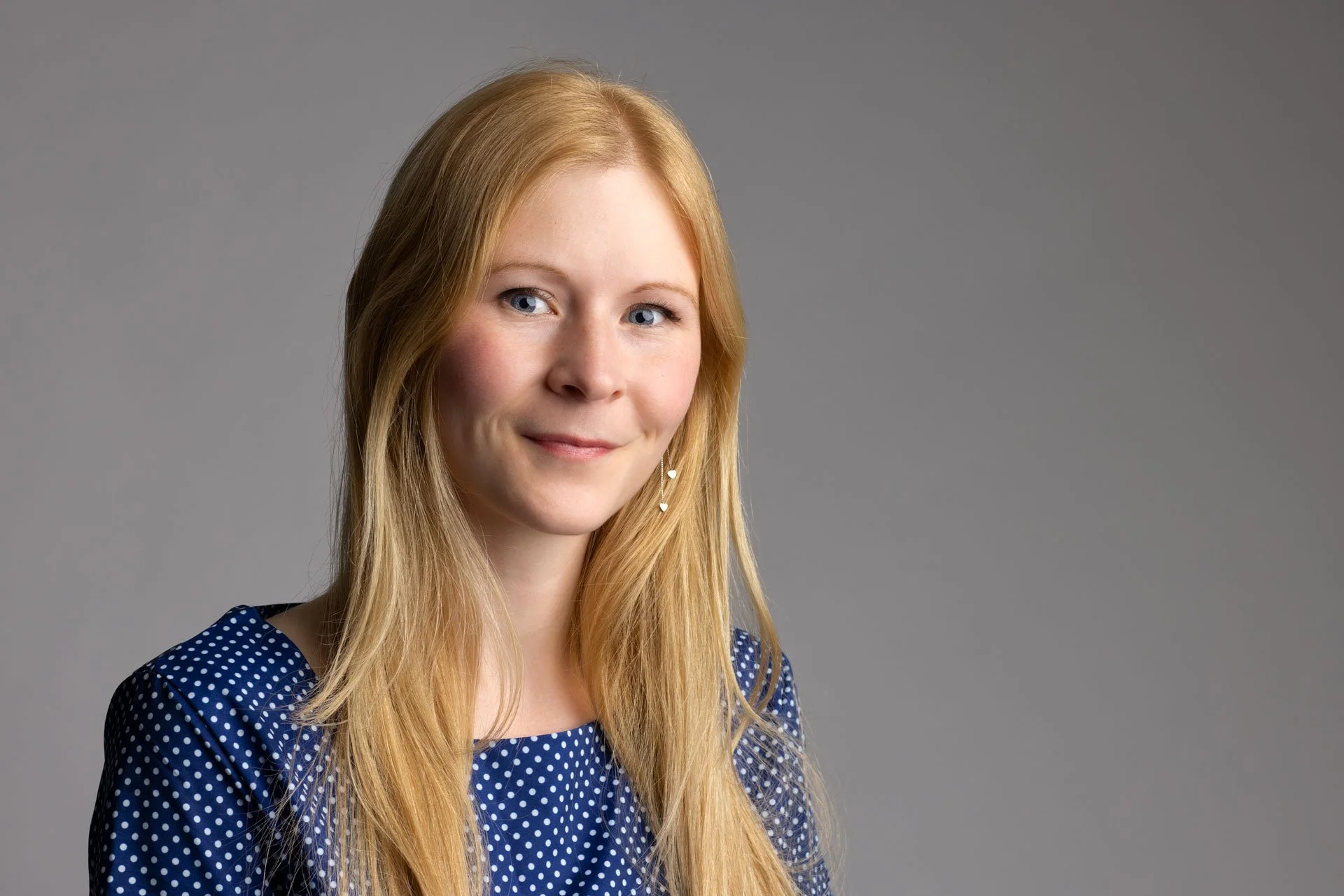 Portræt af en ung kvinde med langt blond hår, blå øjne, iført en blå kjole med hvide prikker, mod en grå baggrund.