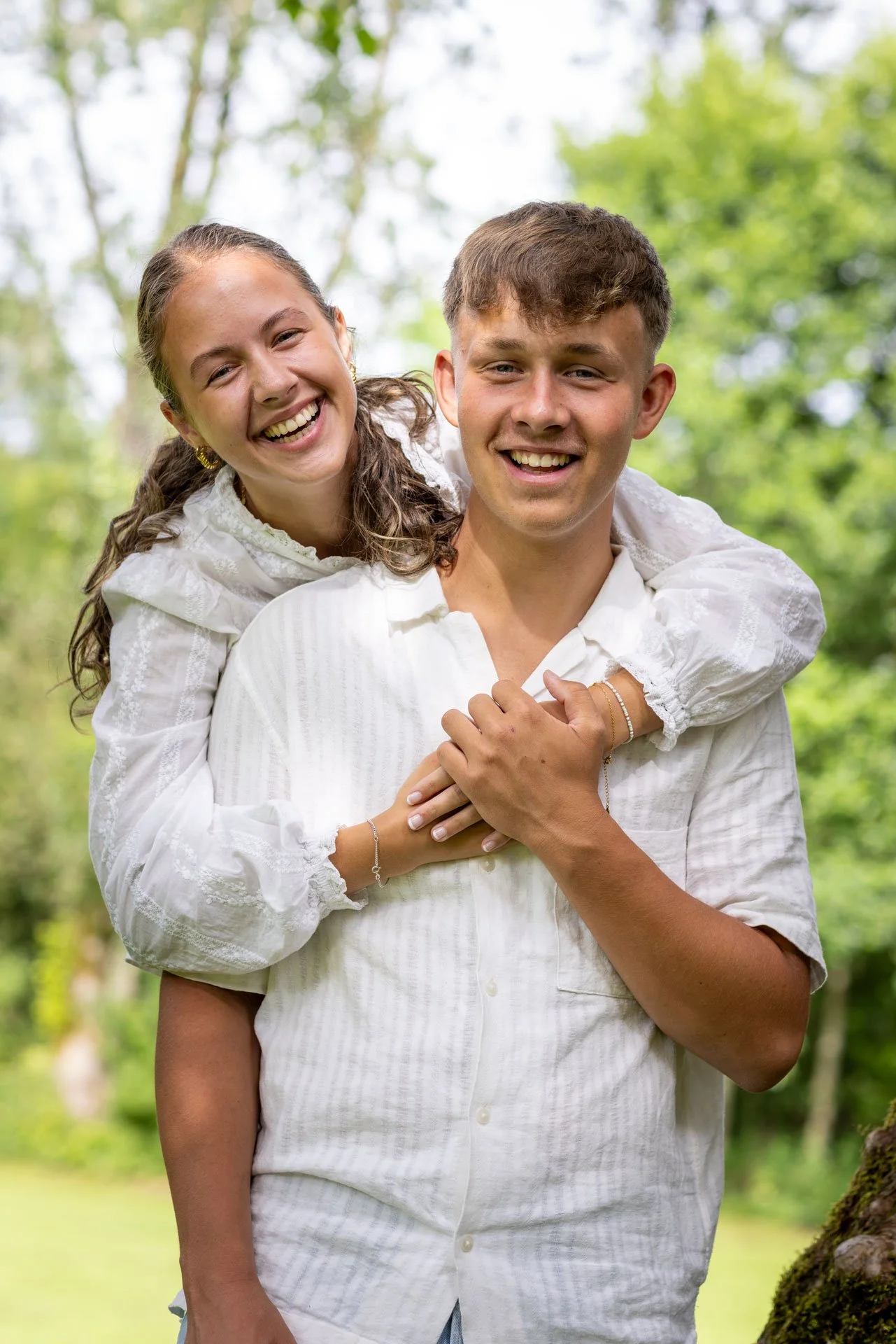 Et smilende par i hvide tøj står udenfor i en grøn skov med lysegrønne blade.