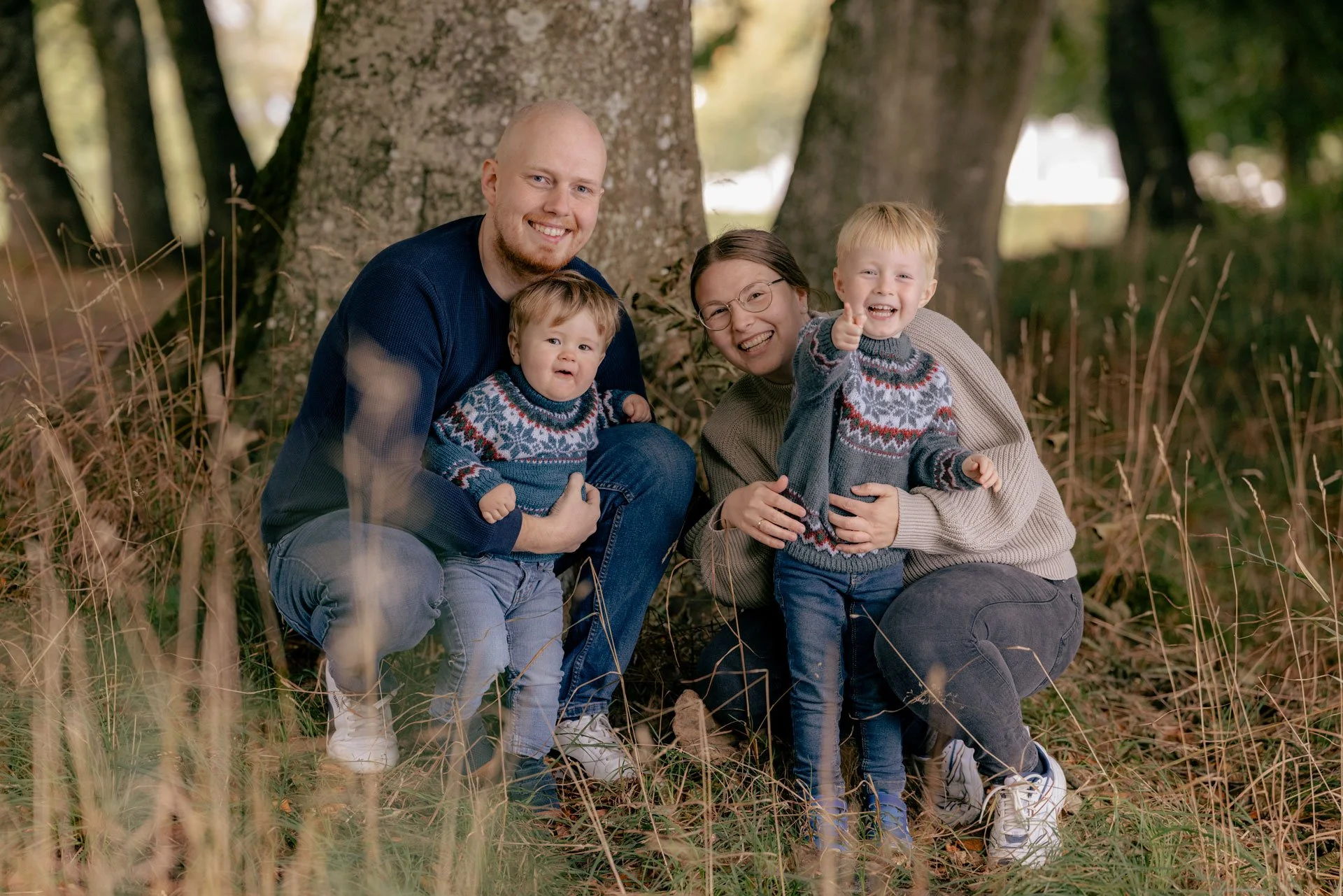 Familie bestående af to voksne og to børn, sidder i en skov med efterårsblade og træer i baggrunden, alle smiler og har på strikkede juletrøjer.